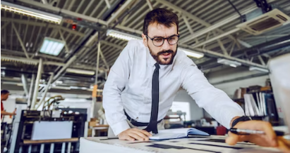 Photo d’un homme en chemise et cravate dans son entreprise en train de 
                réfléchir au-dessus de son bureau avec un stylo à la main et des documents posés.