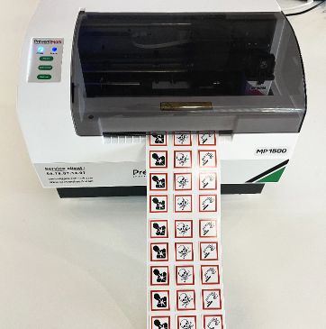 Photo de l’imprimante signalétique MP1500 de
            PREVENTIMARK vue d’au-dessus en train d’imprimer des étiquettes pictogrammes CLP – SGH en format 20 mm x 25 mm. 