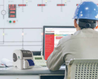 Photo d’un ouvrier muni d’un casque de protection bleu de dos assis sur une chaise et face à
        son bureau sur lequel se trouve son écran d’ordinateur, sur lequel on peut distinguer l’interface du site de PREVENTIMARK et du logiciel MARKPRINT.