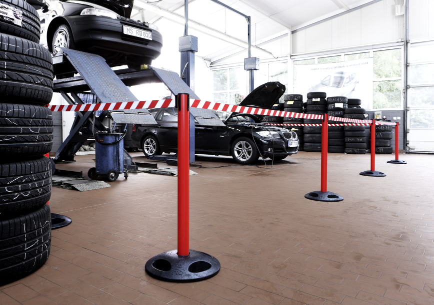 Photo d’un garage automobile avec plusieurs voitures en réparation derrière quatre poteaux reliés et clipsés par des sangles rouges et blanches de façon à barrer le passage vers les voitures. 