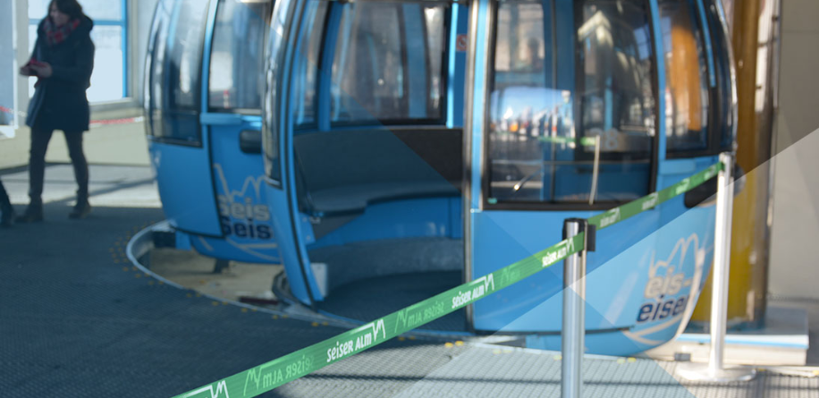 Photo d’un téléphérique bleu où l’accès est balisé par des poteau à sangle portant le logo et les couleurs de la station. 
