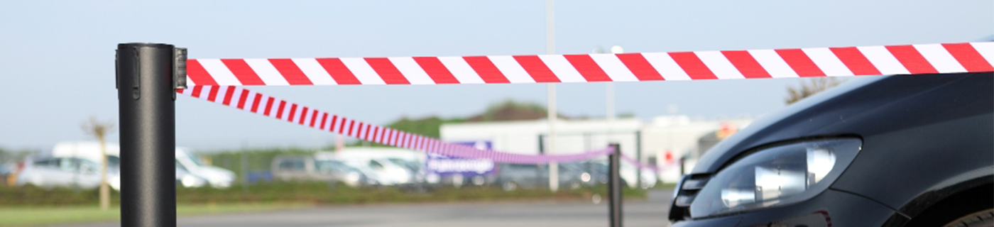 Photo d’une sangle zébrée rouge et blanche tenue par un poteau noir sur un parking en extérieur laissant apercevoir le devant d’une voiture noire. 