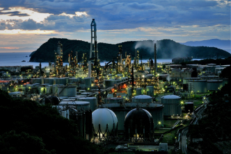 Photo d’un grand site industriel éclairé vu de dehors à la tombée de la nuit avec une colline et la mer en fond